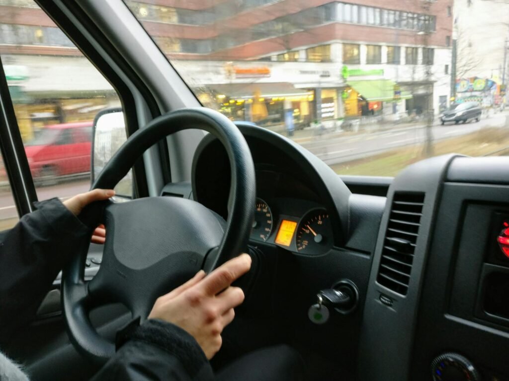 Transporte Ferreira Camarena Toledo Hands on a steering wheel driving a van through an urban area with shops visible outside.